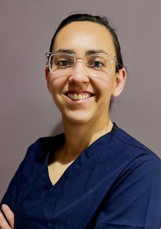 A woman in blue scrubs with glasses smiles.